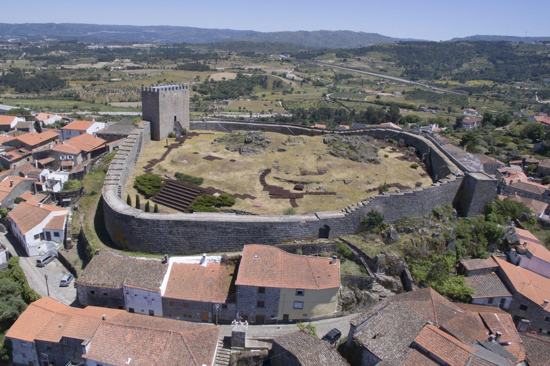 Castelo medieval com torre quadrangular e muralhas de pedra, rodeado por casarões com telhados de telha de barro, inserido numa paisagem montanhosa com campos e vegetação.